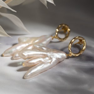 May include: A pair of gold earrings with a unique, organic design. Each earring features a large, irregular-shaped pearl and a gold hoop with a textured finish.