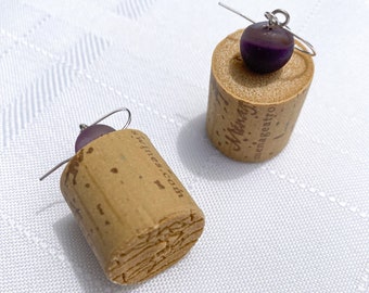 May include: A pair of earrings made from wine corks with purple glass beads. The corks have text on them.