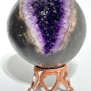 May include: A spherical amethyst geode with a dark grey exterior and a deep purple crystalline interior, featuring a prominent vertical fissure. The geode rests on a metallic rose gold-coloured stand with a decorative heart-shaped cutout design.