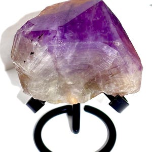 May include: A large, raw amethyst crystal cluster featuring deep purple hues at the top that transition into a translucent, pale base. The crystal is held securely by a black metal display stand with a circular base and three prongs.