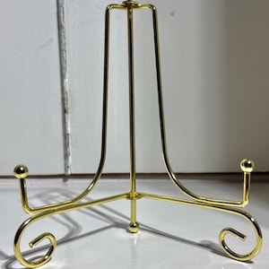 May include: A gold-coloured metal display stand with a three-legged base and a curved top. The stand has a decorative design with rounded accents and is designed to hold a plate or other item.