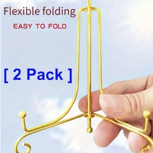 May include: A gold-coloured, flexible folding display stand. The stand is designed to be easy to fold and is sold as a 2-pack. The stand has a three-legged base with decorative ball accents and a vertical support.