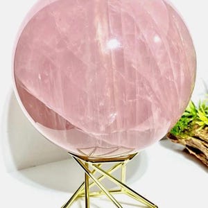 May include: A large, smooth, pale pink rose quartz sphere with visible internal crystalline fractures and natural inclusions. The polished stone orb rests securely on a geometric, gold-toned metal wire stand with a hexagonal base.