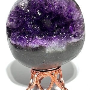 May include: A spherical amethyst geode featuring deep purple, jagged crystal formations within a dark, smooth outer shell. The geode is supported by a metallic, rose-gold coloured stand that has a decorative base with heart-shaped cutouts.