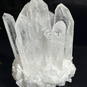 May include: A cluster of clear, translucent quartz crystals featuring multiple pointed terminations rising from a base of smaller, jagged white crystal formations. The structure displays natural facets and internal inclusions with a reflective surface.