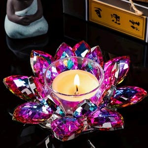 May include: A lotus-shaped candle holder crafted from faceted, iridescent glass petals in vibrant shades of magenta, purple, and blue. The centre features a clear glass cup holding a small, lit tea light candle with a warm yellow flame.