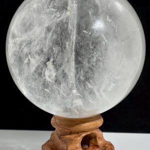 Pode incluir: Uma grande esfera de cristal de quartzo transparente com fraturas internas e inclusões nubladas visíveis, apoiada sobre um suporte de madeira esculpida em tom castanho médio com base circular e quatro pés robustos.