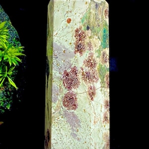 Puede incluir: Un obelisco de cristal rectangular y alto con una parte superior puntiaguda. La piedra presenta una mezcla de colores, incluyendo crema, verde y marrón rojizo, con patrones y texturas intrincados. El obelisco está sobre un fondo oscuro.