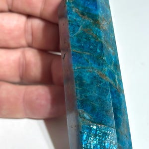Può includere: Una torre di cristallo di apatite blu di forma esagonale, caratterizzata da profonde tonalità azzurre e verde acqua con inclusioni marroni naturali e una texture cristallina riflettente su tutta la superficie della punta in pietra.