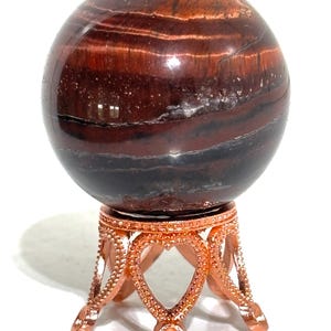 May include: A polished, spherical red tiger's eye gemstone featuring horizontal bands of deep brown, black, and reddish-orange hues. The sphere rests securely on a decorative, metallic copper-coloured stand with intricate heart-shaped cutouts.