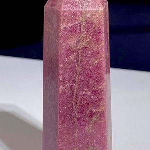 May include: A polished, hexagonal crystal tower featuring a vibrant magenta and deep pink colour palette. The stone displays a textured, crystalline surface with natural inclusions and subtle tan-coloured mineral veins running vertically.