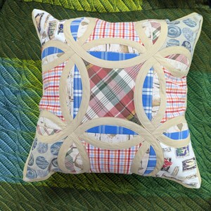 May include: A square throw pillow featuring a double wedding ring quilt pattern. The design incorporates various fabric patches in plaid, checkered, and patterned prints, separated by beige fabric bands that form interlocking circular shapes.