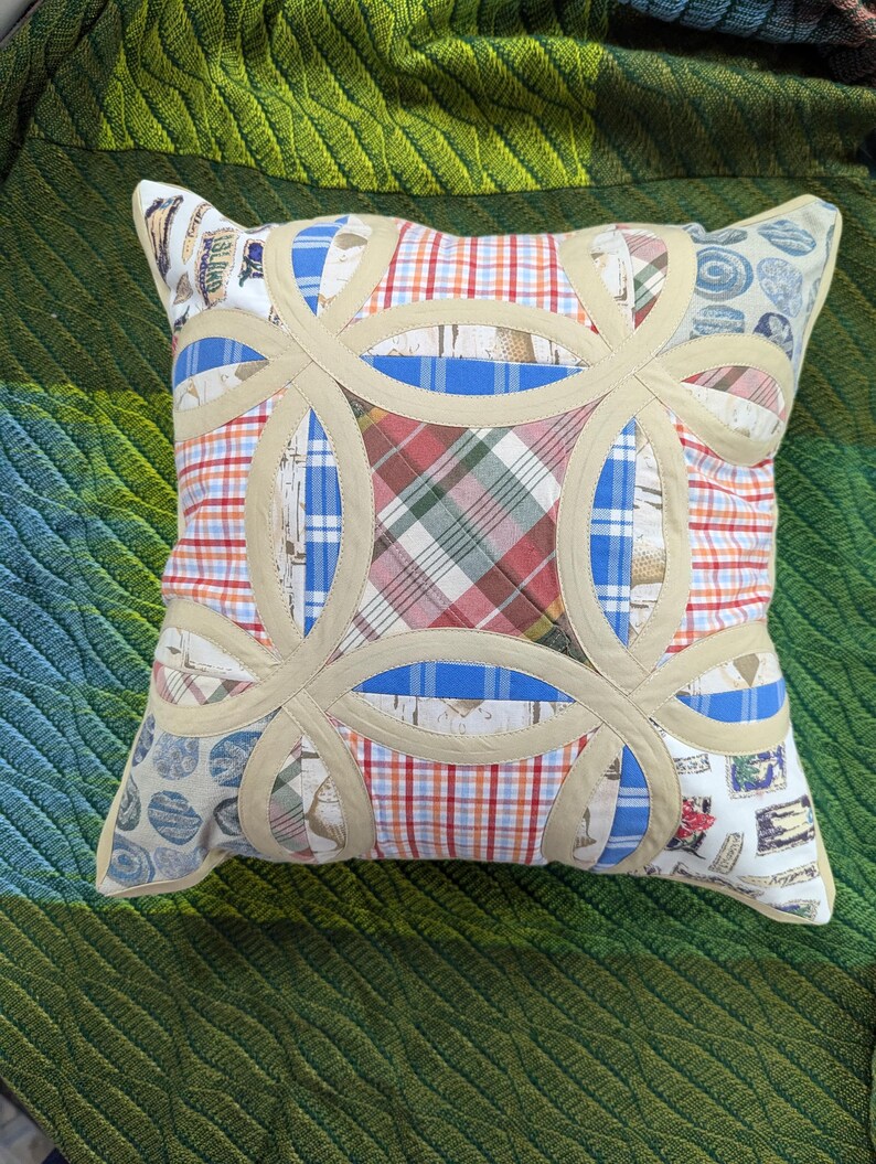 May include: A square throw pillow featuring a double wedding ring quilt pattern. The design incorporates various fabric patches in plaid, checkered, and patterned prints, separated by beige fabric bands that form interlocking circular shapes.