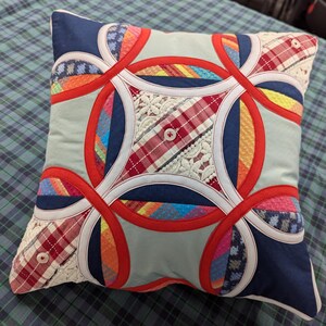 May include: A square throw pillow featuring a double wedding ring quilt pattern. The design incorporates various fabric textures including red, blue, and light blue solids, plaid patterns, and white lace inserts, accented with small white buttons.