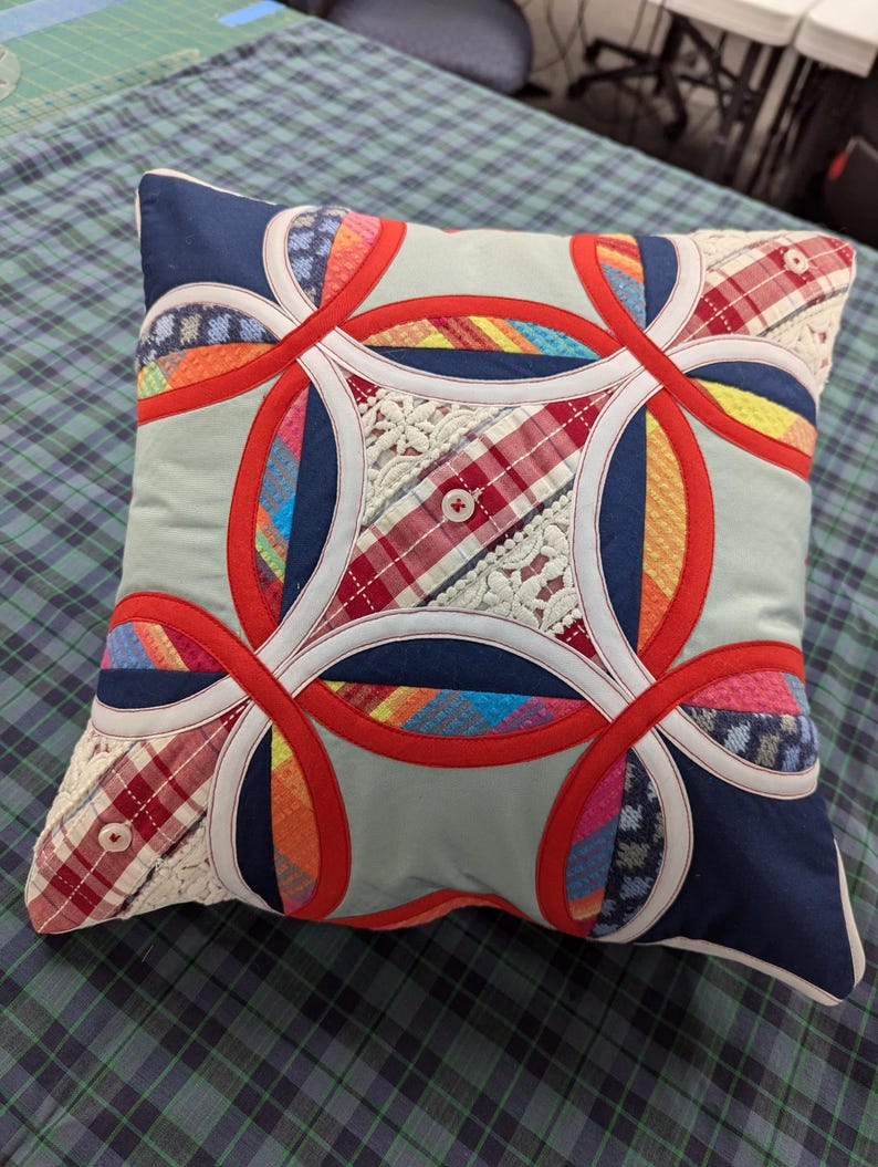 May include: A square throw pillow featuring a double wedding ring quilt pattern. The design incorporates various fabric textures including red, blue, and light blue solids, plaid patterns, and white lace inserts, accented with small white buttons.