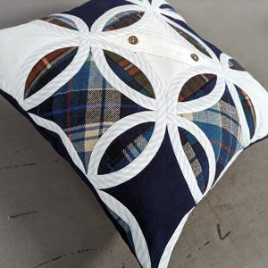 May include: A square throw cushion featuring a patchwork design of navy blue fabric and various plaid patterns in blue, green, and brown. The top surface is accented with white fabric circular overlays and two small brown buttons centred on top.