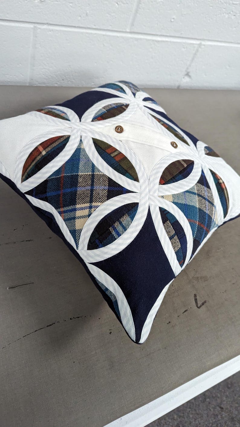 May include: A square throw cushion featuring a patchwork design of navy blue fabric and various plaid patterns in blue, green, and brown. The top surface is accented with white fabric circular overlays and two small brown buttons centred on top.