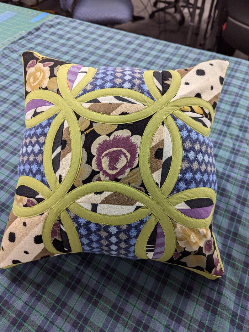May include: A square throw pillow featuring a double wedding ring quilt pattern in lime green fabric. The design incorporates various fabric patches in shades of purple, blue, black, tan, and white, displaying floral, geometric, and polka dot prints.