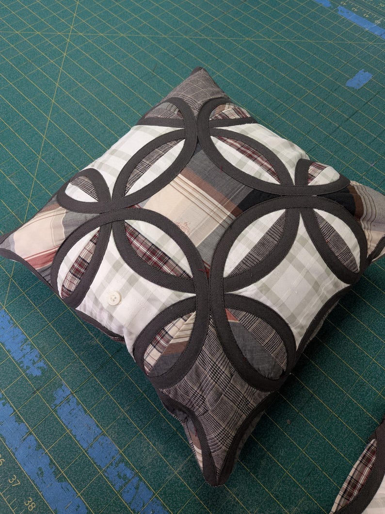 May include: A square throw cushion featuring a patchwork design of various plaid fabrics in shades of grey, white, and brown. The cushion is accented with dark grey fabric strips arranged in overlapping circular patterns and a single white button.