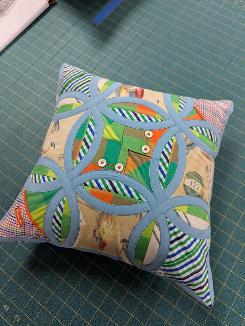 May include: A square throw cushion featuring a double wedding ring quilt pattern in light blue fabric. The design incorporates various patterned fabric patches including stripes, plaids, and beige prints with small illustrations, accented by four white buttons.