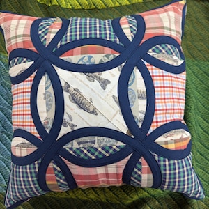 May include: A square throw pillow featuring a double wedding ring quilt pattern in dark navy blue fabric. The pillow is constructed from various fabric patches including red, white, and orange plaid, green and blue gingham, and a cream fish print.