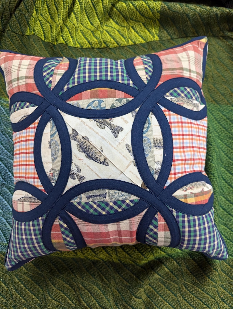 May include: A square throw pillow featuring a double wedding ring quilt pattern in dark navy blue fabric. The pillow is constructed from various fabric patches including red, white, and orange plaid, green and blue gingham, and a cream fish print.