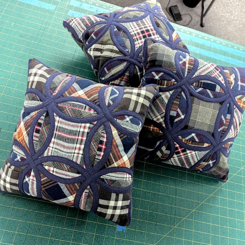 May include: Two square throw pillows featuring a patchwork of various plaid fabrics in shades of blue, grey, white, and brown. Each pillow is decorated with a dark blue fabric overlay arranged in a repeating circular, flower-like pattern.