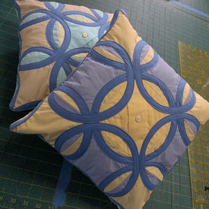 May include: Two square throw pillows featuring a double wedding ring quilt pattern. The pillows are constructed from yellow and blue gingham fabric with solid blue circular appliques and a single white button centered on the front of each pillow.