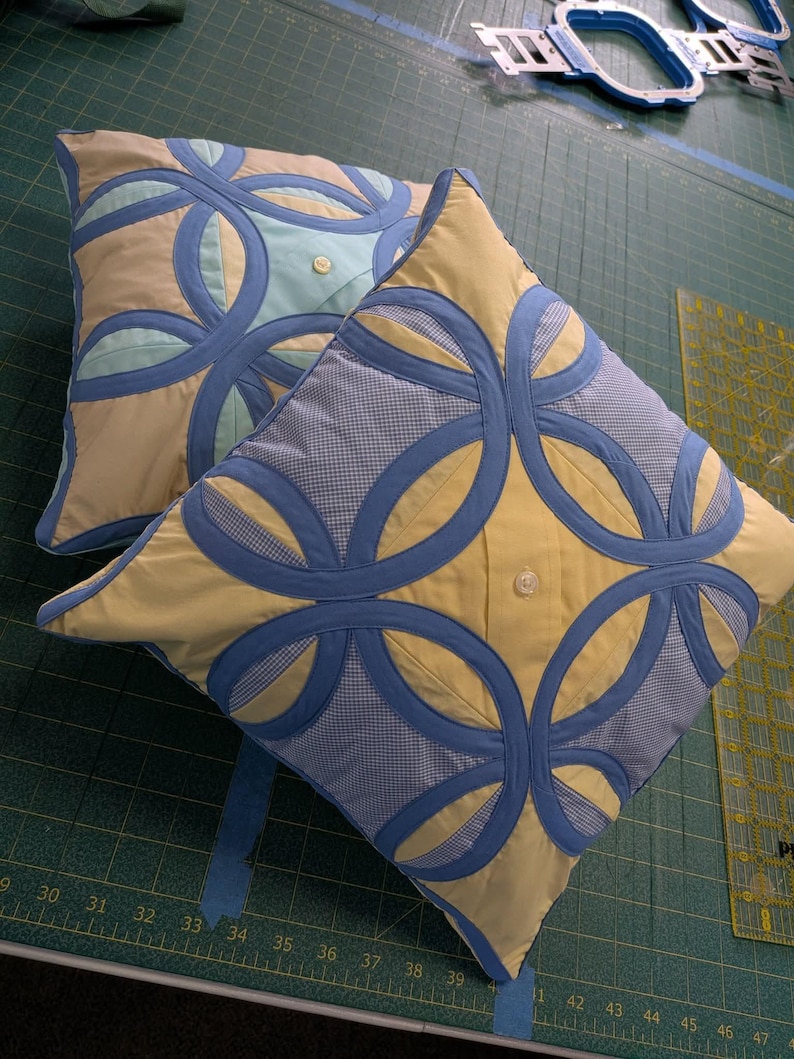 May include: Two square throw pillows featuring a double wedding ring quilt pattern. The pillows are constructed from yellow and blue gingham fabric with solid blue circular appliques and a single white button centred on the front of each pillow.