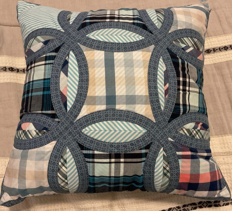 May include: A square throw cushion featuring a double wedding ring quilt pattern. The design is composed of various plaid and striped fabric patches in shades of blue, white, and tan, accented by blue floral patterned fabric borders.