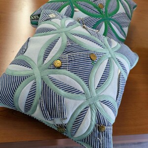 May include: Two square throw pillows crafted from blue and white striped denim fabric. The pillows feature a geometric circular appliqu&eacute; pattern in light green fabric, accented with metallic gold buttons and silver-toned metal hardware.