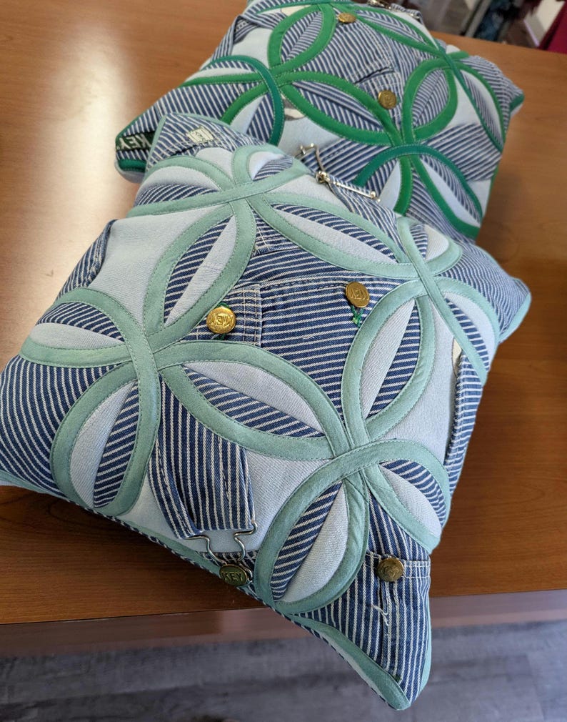 May include: Two square throw pillows crafted from blue and white striped denim fabric. The pillows feature a geometric circular appliqu&eacute; pattern in light green fabric, accented with metallic gold buttons and silver-toned metal hardware.
