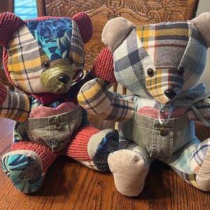May include: Two handmade teddy bears with patchwork fabric bodies. One bear has a red bow tie and red knitted paws. The other bear has a blue scarf. Both bears have denim overalls and heart-shaped pockets. The bears are sitting on a wooden table.