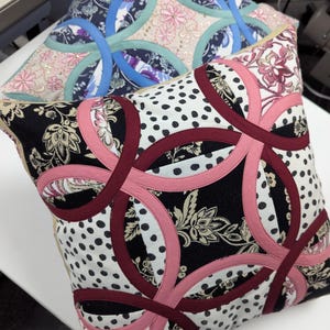 May include: Two square throw pillows featuring a double wedding ring quilt pattern. The front pillow displays black, white, and pink fabric with floral and polka dot prints. The back pillow features blue, teal, and pink fabric with floral patterns.