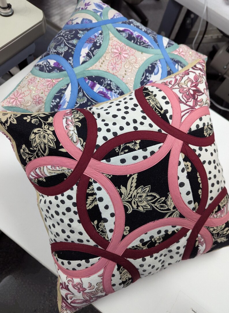 May include: Two square throw pillows featuring a double wedding ring quilt pattern. The front pillow displays black, white, and pink fabric with floral and polka dot prints. The back pillow features blue, teal, and pink fabric with floral patterns.