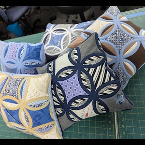 May include: A collection of five square throw cushions featuring a cathedral window quilt pattern. Each cushion displays a unique combination of fabric textures and colours, including shades of blue, tan, black, white, and grey, with stitched circular motifs.