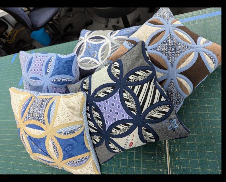 May include: A collection of five square throw cushions featuring a cathedral window quilt pattern. Each cushion displays a unique combination of fabric textures and colours, including shades of blue, tan, black, white, and grey, with stitched circular motifs.
