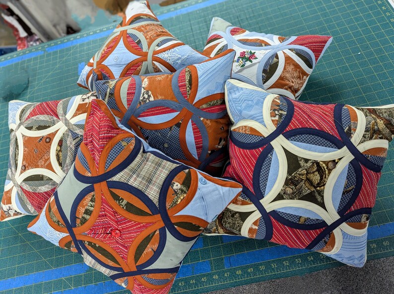 May include: A collection of square throw cushions featuring a double wedding ring quilt pattern. The cushions are constructed from various fabric scraps in shades of red, blue, orange, and brown, with cream-coloured curved bands forming the rings.