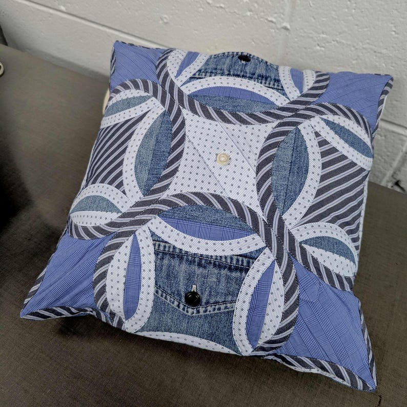 May include: A square throw pillow featuring a patchwork design of blue denim and various blue and white patterned fabrics. The front displays a double wedding ring quilt pattern with a central button and a visible denim pocket with a black button.