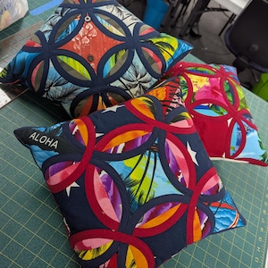 May include: Two square throw pillows featuring a tropical print with palm trees and hibiscus flowers. The pillows are decorated with a dark blue or red circular appliqu&eacute; pattern. One pillow has the word ALOHA embroidered in white on a dark blue strip.