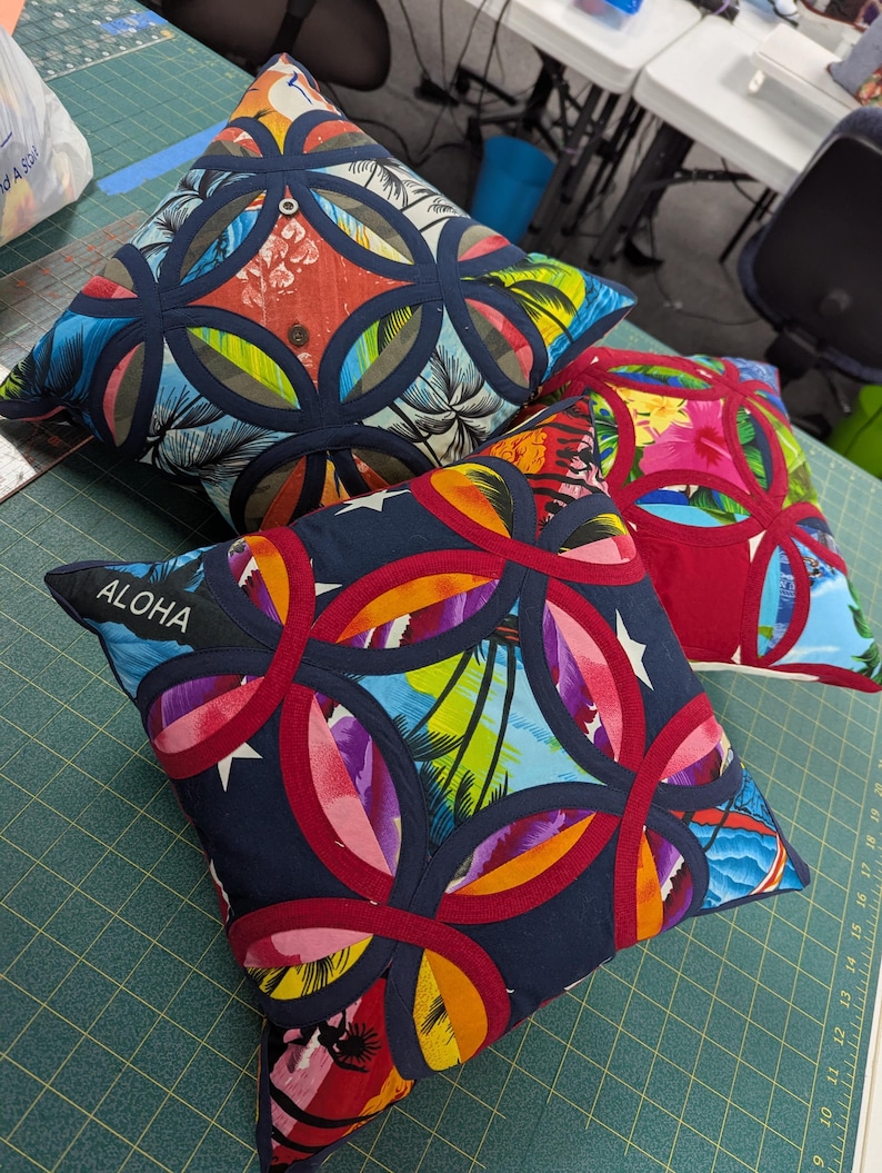 May include: Two square throw pillows featuring a tropical print with palm trees and hibiscus flowers. The pillows are decorated with a dark blue or red circular appliqu&eacute; pattern. One pillow has the word ALOHA embroidered in white on a dark blue strip.