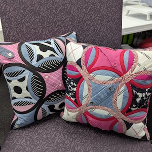 May include: Two square throw pillows featuring a patchwork design of mixed fabrics including denim, pink striped cotton, and black and white patterns. The pillows are constructed with circular motifs, visible buttons, and stitched seams throughout.