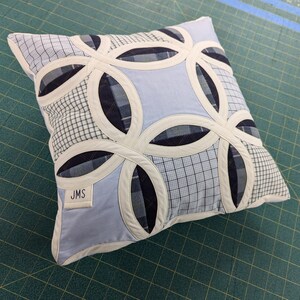 May include: A square throw cushion featuring a double wedding ring quilt pattern made from various blue and white plaid and solid fabric patches. The design includes cream-coloured fabric borders and a small white label with the letters JMS printed on it.