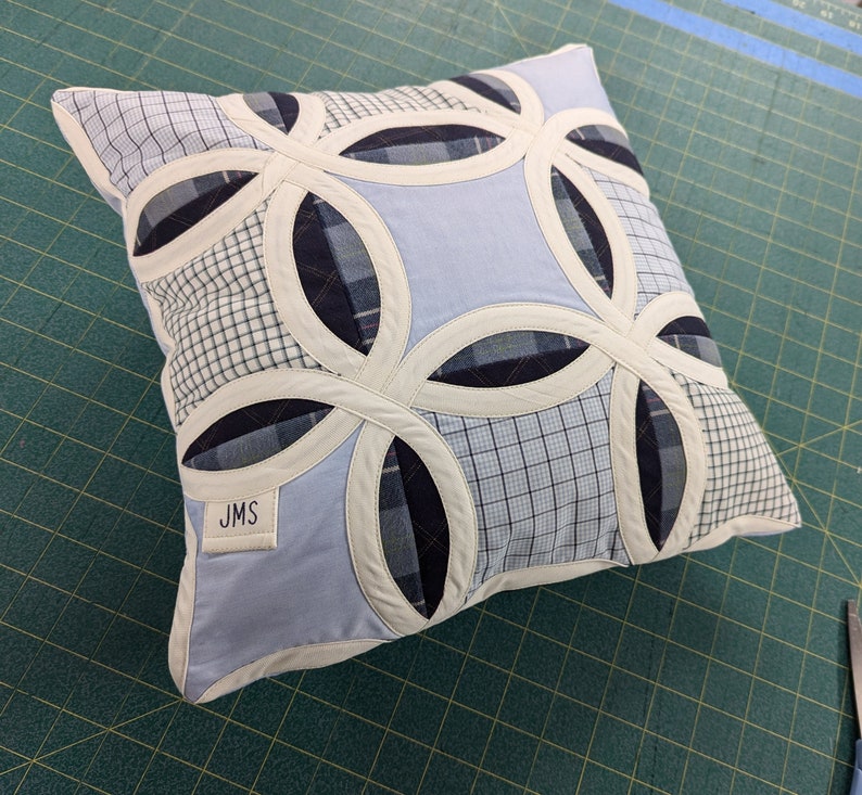 May include: A square throw cushion featuring a double wedding ring quilt pattern made from various blue and white plaid and solid fabric patches. The design includes cream-coloured fabric borders and a small white label with the letters JMS printed on it.