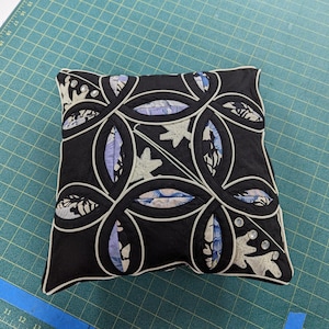 May include: A square decorative cushion featuring a black fabric base with a white piped interlocking circular pattern. The design includes inset panels of light blue and white floral fabric, accented with small clear circular embellishments.