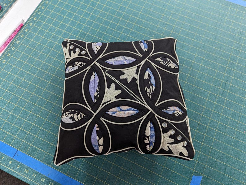 May include: A square decorative cushion featuring a black fabric base with a white piped interlocking circular pattern. The design includes inset panels of light blue and white floral fabric, accented with small clear circular embellishments.
