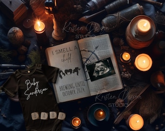 May include: A black onesie with the text "Baby Sanderson" and the word "BABY" spelled out with wooden blocks. The onesie is lying on a dark blue surface with a book open to a page that reads "I smell a child... Brewing until October 2024". There are candles and other decorative items around the book.
