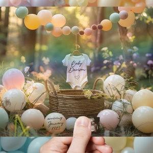 May include: A white onesie with the text "Baby Davison" and a heart design. The onesie is hanging on a clothesline with a string of pastel balloons in the background. The balloons are pink, peach, yellow, and blue. There is a basket of pastel colored balloons in the foreground.