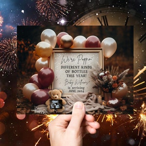 May include: A baby announcement card with a rustic theme. The card features a sign that says "We're Poppin' Different Kinds of Bottles This Year! Baby Williams is arriving June 2024". The sign is surrounded by balloons and a floral arrangement.