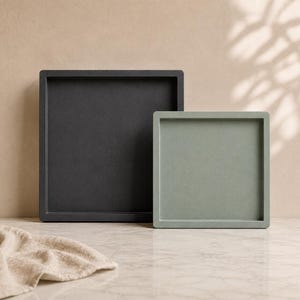 May include: Two square trays, one dark gray and one light green, are displayed. The trays have a minimalist design with a raised edge. A beige cloth is draped in the foreground. The background is a neutral color with shadows.